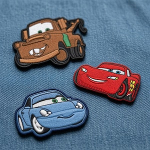 May include: Three embroidered fabric patches featuring cartoon car characters. One is a brown tow truck with a wide grin, one is a red race car with a smiling expression, and one is a light blue sports car with a friendly face and green eyes.