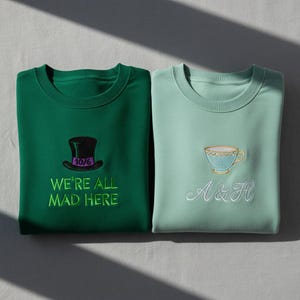 Op de afbeelding: Twee crewneck sweatshirts. Eén is smaragdgroen met een hoge hoed en de tekst "WE'RE ALL MAD HERE". De andere is mintgroen met een theekopje en de letters "A & H".