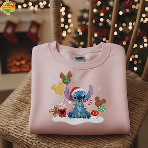 K&ouml;nnte beinhalten: Ein hellrosa Sweatshirt mit einem gestickten Weihnachtsdesign. Das Design zeigt eine blaue Figur mit Weihnachtsm&uuml;tze, umgeben von Lichterketten, Lebkuchen und Mickey Mouse-Ballons und Leckereien. Das Sweatshirt liegt gefaltet auf einem Korbstuhl.