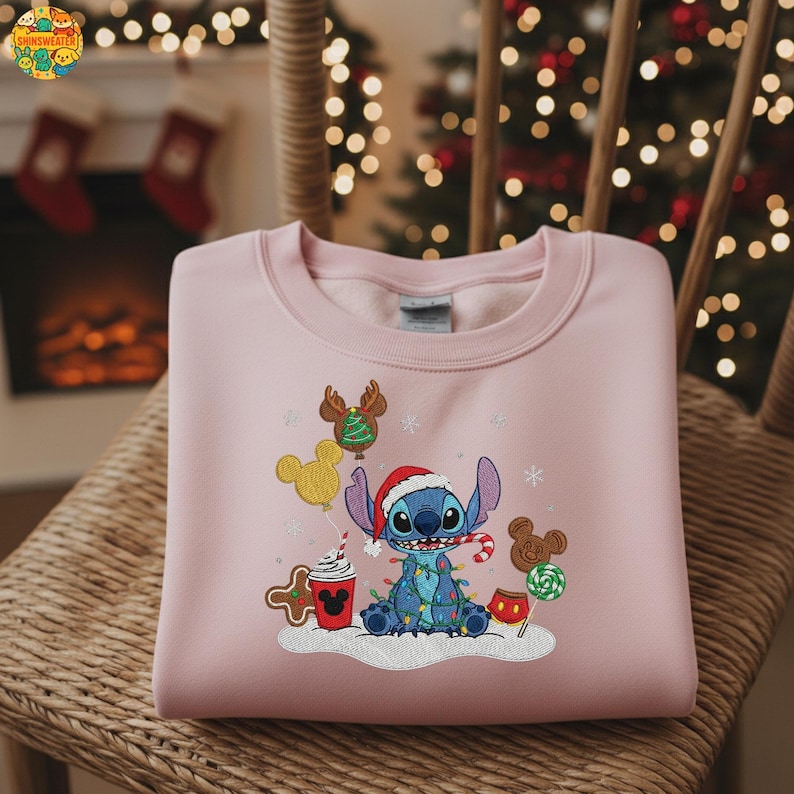 K&ouml;nnte beinhalten: Ein hellrosa Sweatshirt mit einem gestickten Weihnachtsdesign. Das Design zeigt eine blaue Figur mit Weihnachtsm&uuml;tze, umgeben von Lichterketten, Lebkuchen und Mickey Mouse-Ballons und Leckereien. Das Sweatshirt liegt gefaltet auf einem Korbstuhl.