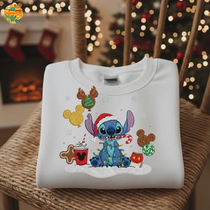K&ouml;nnte beinhalten: Wei&szlig;es Sweatshirt mit einem blauen Cartoon-Charakter, der einen Weihnachtsmannhut tr&auml;gt und von Lichterketten umgeben ist. Das Design umfasst Lebkuchen und eine Kaffeetasse. Das Sweatshirt befindet sich auf einem Korbstuhl.