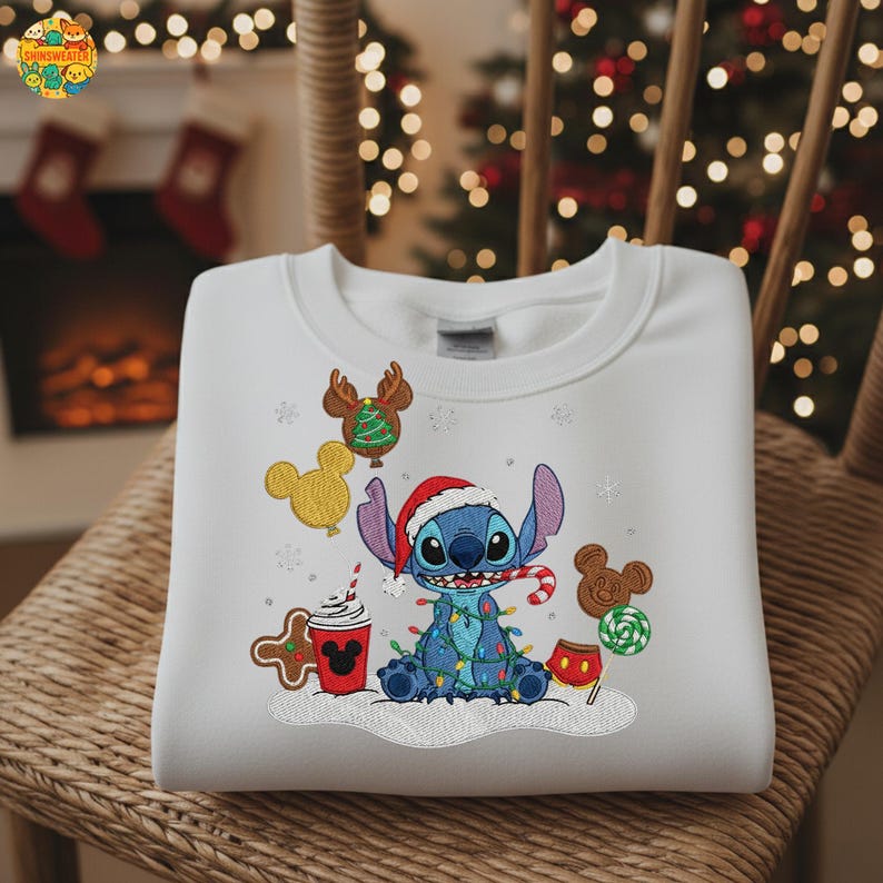 K&ouml;nnte beinhalten: Wei&szlig;es Sweatshirt mit einem blauen Cartoon-Charakter, der einen Weihnachtsmannhut tr&auml;gt und von Lichterketten umgeben ist. Das Design umfasst Lebkuchen und eine Kaffeetasse. Das Sweatshirt befindet sich auf einem Korbstuhl.