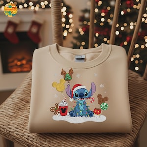 K&ouml;nnte beinhalten: Beiges Sweatshirt mit gesticktem Stitch-Charakter mit Weihnachtsm&uuml;tze, umgeben von Weihnachtsdekorationen. Stitch ist mit Lichterketten umwickelt und h&auml;lt eine Zuckerstange. Weitere Elemente sind Mickey Mouse-Ballons, Lebkuchenmann und eine Tasse Kakao.