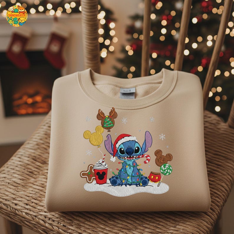 K&ouml;nnte beinhalten: Beiges Sweatshirt mit gesticktem Stitch-Charakter mit Weihnachtsm&uuml;tze, umgeben von Weihnachtsdekorationen. Stitch ist mit Lichterketten umwickelt und h&auml;lt eine Zuckerstange. Weitere Elemente sind Mickey Mouse-Ballons, Lebkuchenmann und eine Tasse Kakao.
