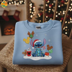 K&ouml;nnte beinhalten: Hellblaues Sweatshirt mit festlichem Stickdesign. Das Design zeigt eine blaue Cartoonfigur mit Weihnachtsm&uuml;tze, umgeben von Weihnachtsdekorationen, Schneeflocken und Luftballons. Das Sweatshirt wird auf einem Korbstuhl pr&auml;sentiert.