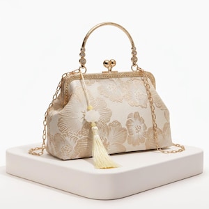 Peut inclure: Sac à main ivoire à motifs floraux avec une monture, une poignée et une bandoulière en chaîne dorée. Le sac est orné d'un pompon décoratif et d'une fermeture à fermoir. Le motif floral est d'une teinte ivoire plus claire.