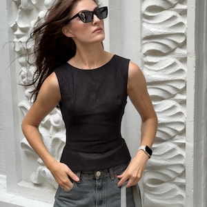 Peut inclure: Un haut noir sans manches et un jean gris. La personne porte des lunettes de soleil noires et une montre. Un sac à main gris est tenu à la main. Le fond est un mur blanc avec des moulures décoratives.