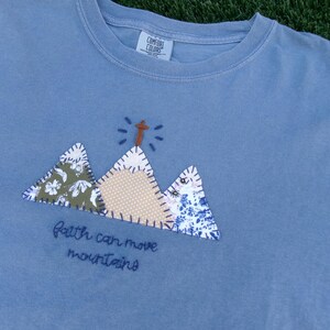 May include: A dusty blue crew neck t-shirt featuring an embroidered applique design of three mountains with patterned fabric patches. The center mountain is topped with a cross and the text "faith can move mountains" is stitched below in dark blue.