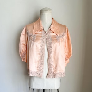May include: A peach-colored satin jacket with short sleeves and a collared neckline. The jacket features delicate lace trim along the front opening and sleeve cuffs. The garment is displayed on a vintage dress form.