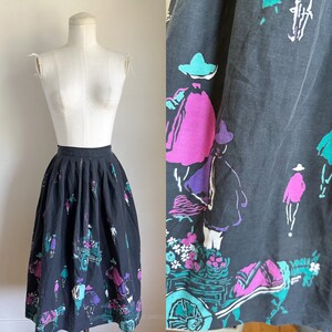 May include: A black midi skirt with a colorful print of people and a wagon. The print features shades of pink, purple, teal, and white.