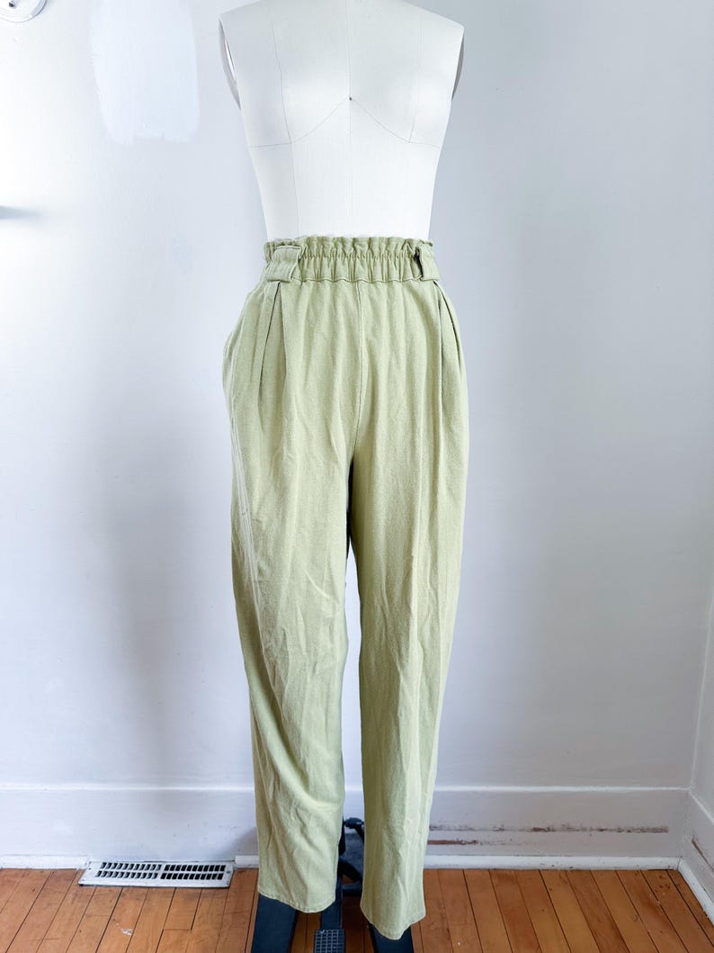 May include: Light green high-waisted pants with a gathered waistband and pleats. The pants are displayed on a white mannequin. The fabric appears to be a lightweight material, suitable for warmer weather.