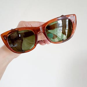 May include: Vintage cat-eye sunglasses with a translucent amber frame and green-tinted lenses. The sunglasses have a classic design with a slightly curved shape and small decorative details on the frame.