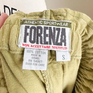 May include: Close-up of olive green shorts with an elastic waistband. A tag reads "ATHENTIC SPORTWEAR FORENZA" in black and white. The tag also states "100% Cotton, Made in Israel, RN 54867, Over for Care" and the size is "S".