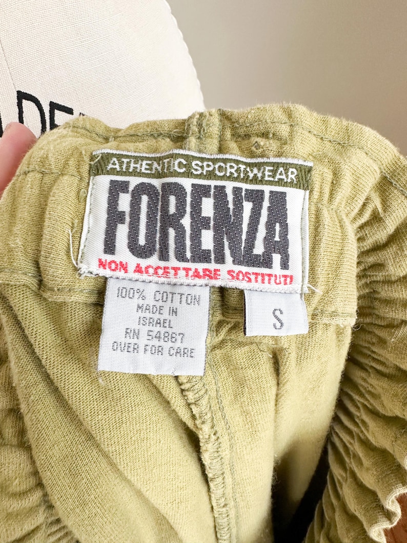 May include: Close-up of olive green shorts with an elastic waistband. A tag reads "ATHENTIC SPORTWEAR FORENZA" in black and white. The tag also states "100% Cotton, Made in Israel, RN 54867, Over for Care" and the size is "S".
