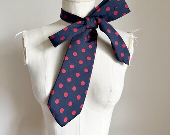 May include: A navy blue scarf with a repeating pattern of red polka dots is tied around a mannequin's neck. The scarf is made of a smooth fabric and is knotted at the center, with the ends hanging down. The scarf is a classic accessory.