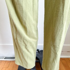 May include: Close-up of light green pants. The fabric has a textured appearance. The pants are straight-legged and appear to be made of a soft material. The pants are displayed on a black stand, with a wooden floor in the background.