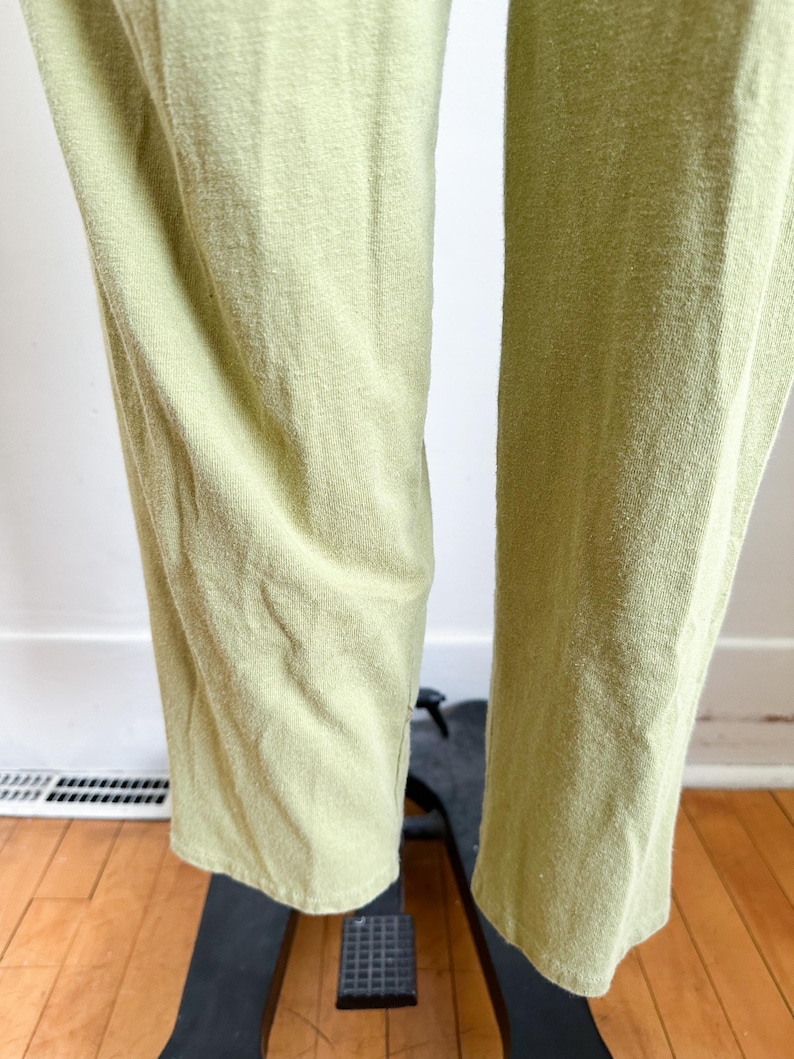 May include: Close-up of light green pants. The fabric has a textured appearance. The pants are straight-legged and appear to be made of a soft material. The pants are displayed on a black stand, with a wooden floor in the background.