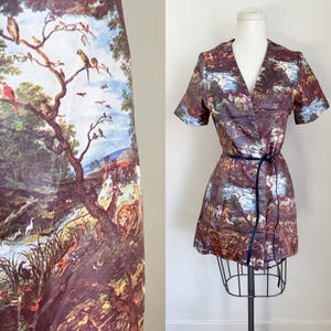 May include: A short-sleeved wrap dress with a landscape print featuring trees, birds, and figures. The dress is cinched at the waist with a dark blue belt. The fabric has a brown, blue, and green color palette.
