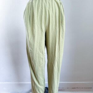 May include: Light green high-waisted pants with a gathered waistband and pleats. The pants are made of a soft, textured fabric and have a relaxed fit. The pants are displayed on a mannequin against a white wall and wooden floor.