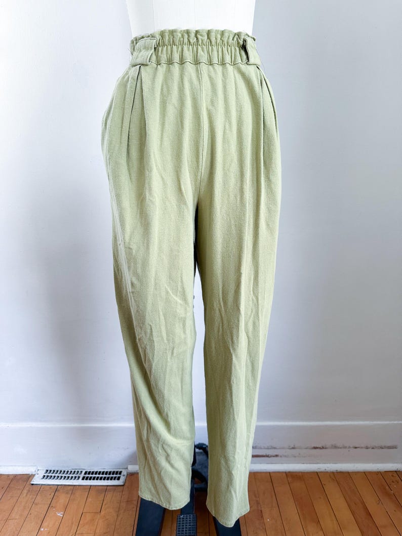 May include: Light green high-waisted pants with a gathered waistband and pleats. The pants are made of a soft, textured fabric and have a relaxed fit. The pants are displayed on a mannequin against a white wall and wooden floor.