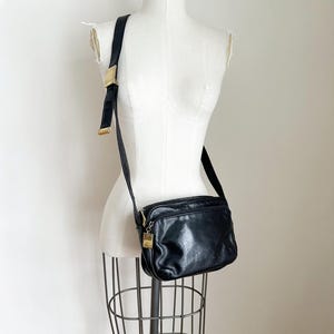 Puede incluir: Bolso bandolera negro con forma rectangular y correa larga ajustable. El bolso presenta una hebilla y cremallera doradas. El bolso se exhibe en un maniquí blanco.