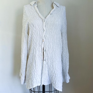 May include: White long-sleeve shirt with a textured, crinkled fabric. The shirt has a collared neckline and a button-down front. The sleeves are long, and the shirt is displayed on a mannequin. The overall style is casual and relaxed.