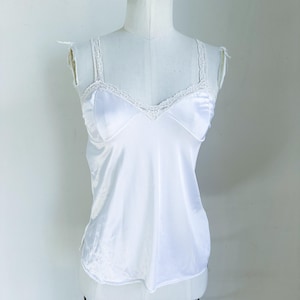 Peut inclure: Un caraco en satin blanc avec une délicate bordure en dentelle sur l'encolure et les bretelles. Le caraco est présenté sur un mannequin, mettant en valeur son tissu lisse et son design classique. Le vêtement a un décolleté en V et des bretelles réglables.