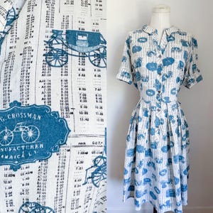 May include: A vintage white dress with a blue print featuring carriages, clocks, and text. The dress has short sleeves, a collared neckline, and a button-down front. The fabric has a pattern of vertical lines and various shapes.