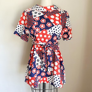 May include: A short-sleeved dress featuring a patchwork pattern of red, white, and navy blue colors. The design includes floral motifs and polka dots throughout the fabric. A matching fabric belt ties at the waist, and the dress has a high collar.