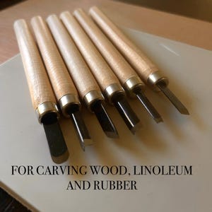 Pode incluir: Conjunto de seis ferramentas para entalhar madeira, com cabos de madeira e lâminas de metal. As ferramentas estão dispostas diagonalmente, mostrando suas bordas afiadas. Texto na parte inferior: FOR CARVING WOOD, LINOLEUM AND RUBBER.