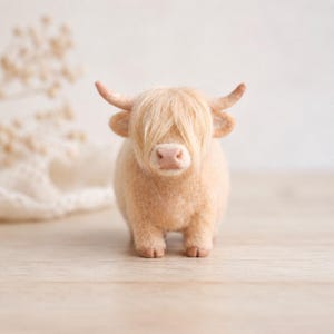 Könnte beinhalten: Eine nadelgefilzte Highland-Rind-Figur in hellem Cremeton. Die kleine Tierfigur besitzt langes, zotteliges Haar, das die Augen bedeckt, zwei geschwungene Hörner, eine zartrosa Nase und vier kurze Beine, die auf einer ebenen Fläche stehen.