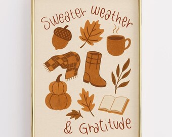 May include: A framed print with a gold border features fall-themed illustrations. The artwork includes a coffee cup, leaves, a scarf, a boot, a pumpkin, and an acorn. The words "Sweater Weather & Gratitude" are written in brown script.