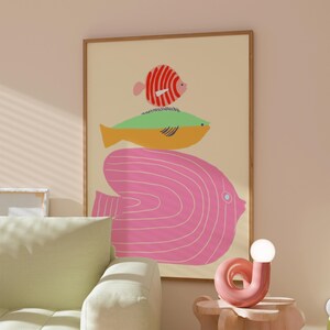 May include: A framed art print depicting a stack of stylized fish in vibrant colours. The artwork showcases a large pink fish, a yellow and green fish, and a small red and white fish, set against a light beige backdrop. The frame is wooden.