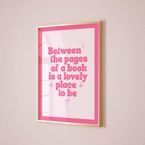 May include: A framed print with a pink border and gold frame. The print features the text "Between the pages of a book is a lovely place to be" in pink, stylized font. The background is white.