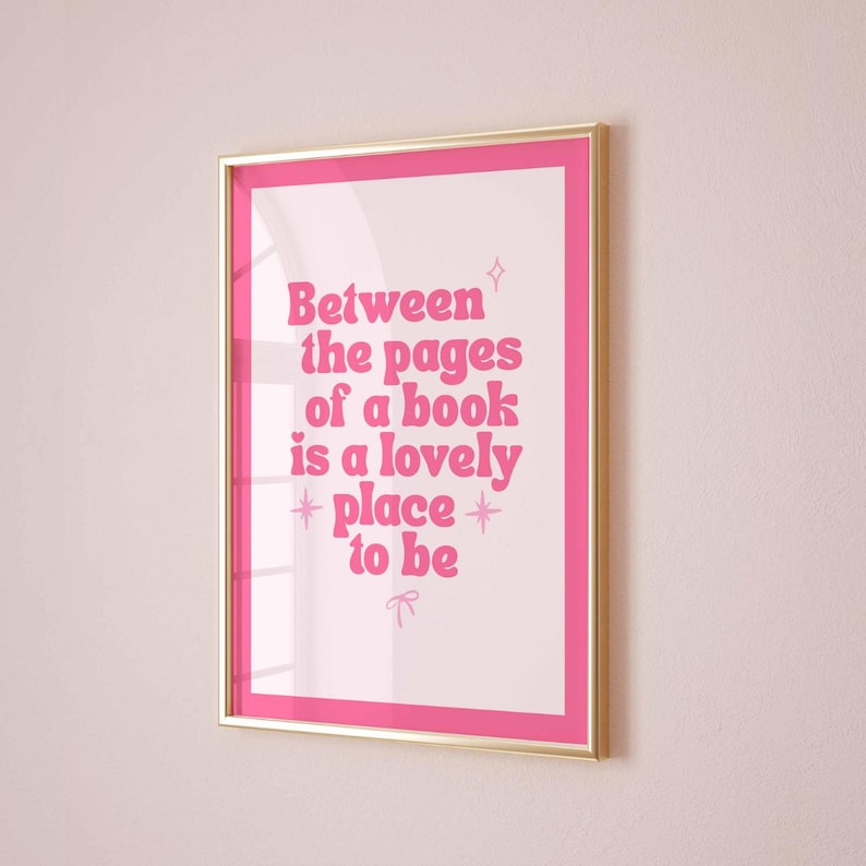 May include: A framed print with a pink border and gold frame. The print features the text "Between the pages of a book is a lovely place to be" in pink, stylized font. The background is white.