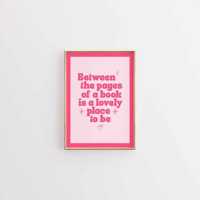May include: A framed print with a pink background and gold frame. The print features the text "Between the pages of a book is a lovely place to be" in pink, stylized lettering. A decorative bow is at the bottom.