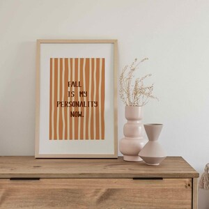 Peut inclure: Une affiche encadr&eacute;e avec le texte "FALL IS MY PERSONALITY NOW" en lettres marron sur un fond de rayures orange verticales. Deux vases d&eacute;coratifs et des plantes s&eacute;ch&eacute;es sont pos&eacute;s sur une surface en bois.