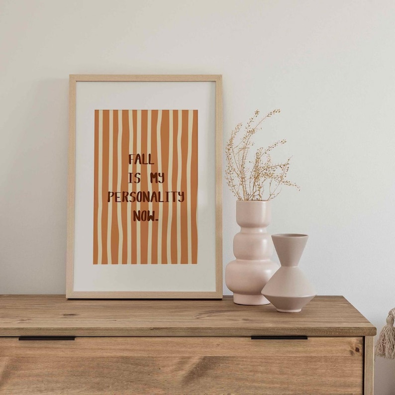 Peut inclure: Une affiche encadr&eacute;e avec le texte "FALL IS MY PERSONALITY NOW" en lettres marron sur un fond de rayures orange verticales. Deux vases d&eacute;coratifs et des plantes s&eacute;ch&eacute;es sont pos&eacute;s sur une surface en bois.