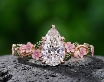 May include: A pear-shaped clear gemstone ring set in a gold-toned band. The band features decorative enamel details including small pink flowers and green leaves that extend along the sides of the central stone setting in a vine-like pattern.