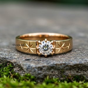 May include: A gold ring featuring a central, clear, round-cut diamond. The band is decorated with starburst engravings. The ring is set on a grey stone surface with green moss in the foreground.
