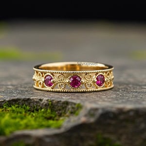 Puede incluir: Un anillo de banda de oro con tres piedras preciosas rojas redondas y pequeñas piedras claras. El anillo tiene un diseño de filigrana ornamentado. El anillo está sobre una superficie de piedra con musgo verde.