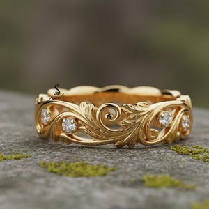 以下が含まれることがあります： 精巧なフローラルデザインのゴールドカラーのリング。リングには、詳細なパターンに小さな輝く宝石がセットされています。リングは、質感のある灰色の表面に表示されています。