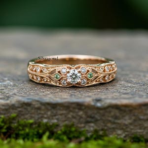 May include: Rose gold ring with a central diamond and smaller diamonds and green gemstones. The ring features an intricate filigree design. The ring is set on a stone surface with a blurred green background.