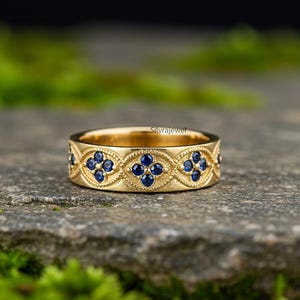 May include: A gold-colored ring with a repeating floral pattern of small, dark blue gemstones. The ring is set on a textured, gray stone surface, with a blurred green background. The ring is engraved with the word "Silvrajewel".