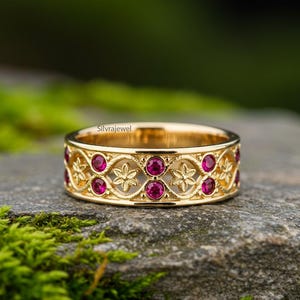 May include: A gold-colored ring with a floral and geometric design, set with multiple round, red gemstones. The ring is displayed on a gray stone surface with green moss in the foreground. The brand name "Silvrajewel" is visible.