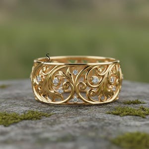 以下が含まれることがあります： 複雑な花の透かし彫りデザインが施された、幅広のゴールドカラーのリング。 小さな透明な宝石が透かし模様にセットされています。 リングは灰色の石の表面に展示されています。