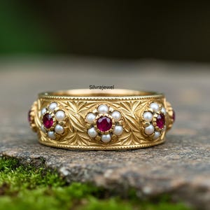 May include: A gold-colored ring with a floral design. The ring features small, round, white pearls and red gemstones set in a repeating pattern. The band has an engraved leaf pattern.