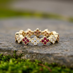 Op de afbeelding: Een goudkleurige ring met een bloemmotief. De ring heeft afwisselende clusters van rode en witte edelstenen. De edelstenen zijn in een herhalend patroon rond de band geplaatst. De ring wordt getoond op een stenen oppervlak.