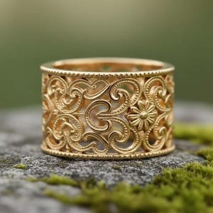 以下が含まれることがあります： 金色のリング。繊細な透かし彫りのデザインで、花柄とスクロール模様が施されています。リングは円筒形で、緑色の苔が生えた石の上に置かれています。リングの精巧なデザインは、ぼやけた緑色の背景の中で際立っています。