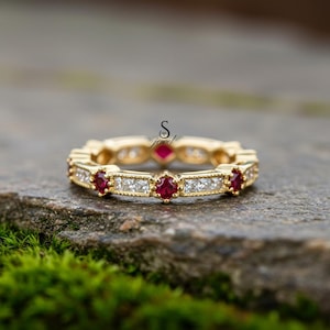 May include: A gold ring with a repeating pattern of red gemstones and square-cut white gemstones. The ring is set on a natural stone surface with green moss in the foreground. The ring has the letter "S" and the word "jewel" in the image.